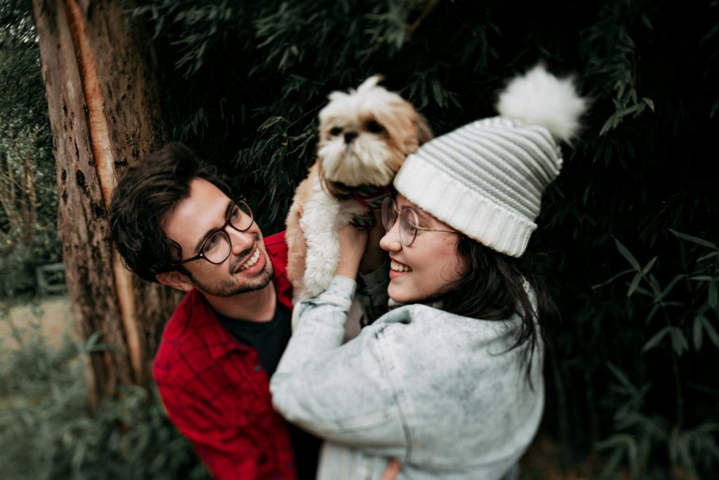 A joyful couple smiling and holding a Shih Tzu dog outdoors in a lush park setting.