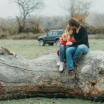 A couple embraces tenderly on a large fallen tree, set against a serene natural landscape.